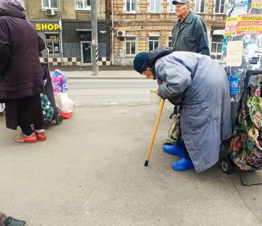 «Всю пенсию отдаю, вдвоём выживаем»: как живут люди, каждый день ждущие на остановке в Ростове бесплатного обеда Сгорбленная старушка стоит, облокотившись о столб. Сумка рядом. Парни кладут туда еду. //Фото Инна Лебедева